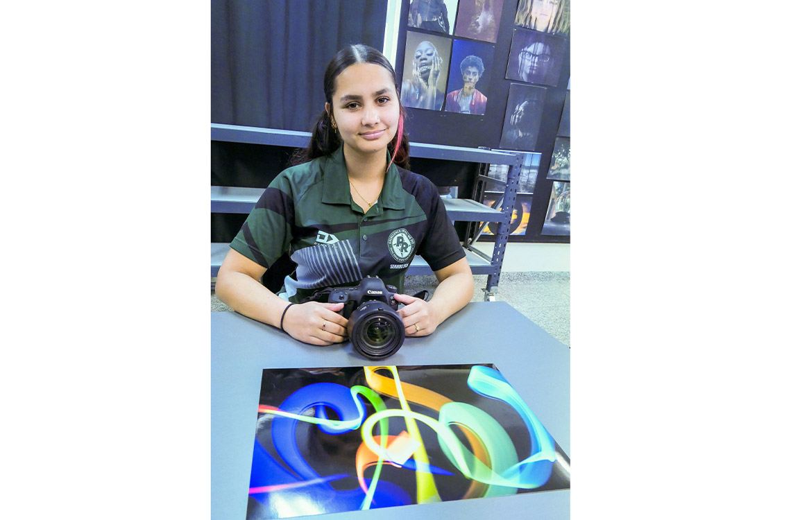 High school student Amali sits with her camera in front of her photographic artwork