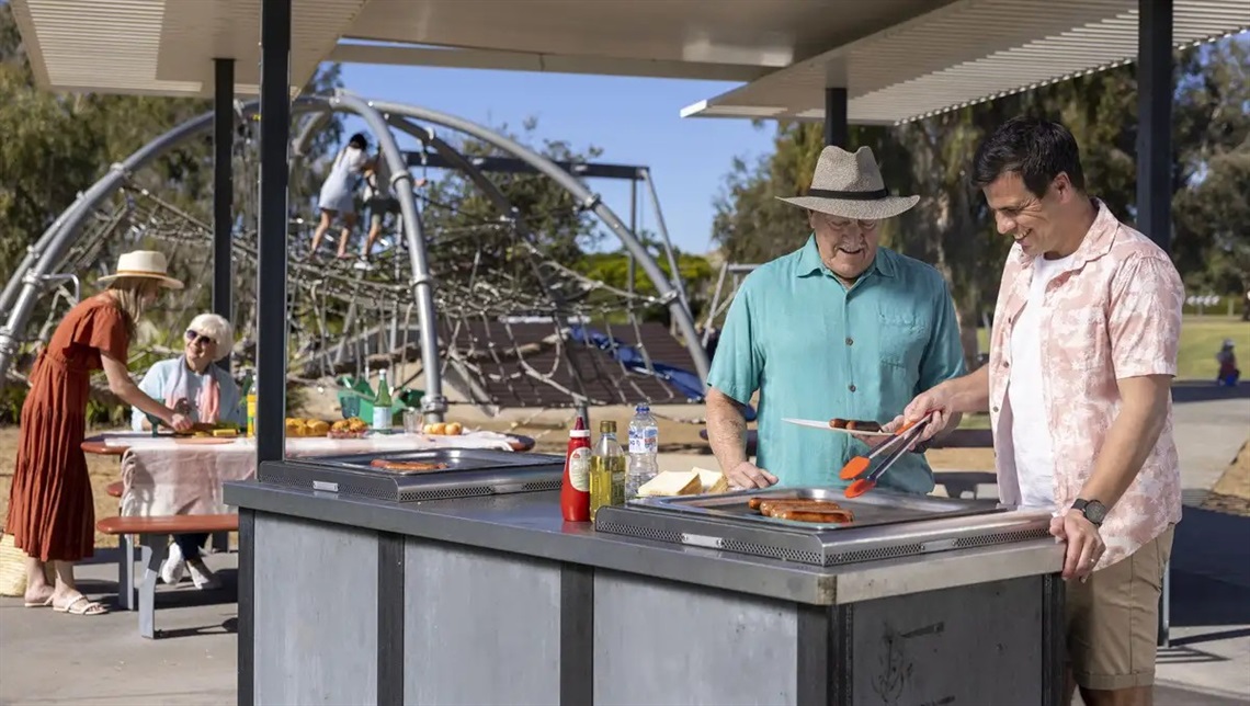 Two fathers standing at a barbeque in a park cooking sausages