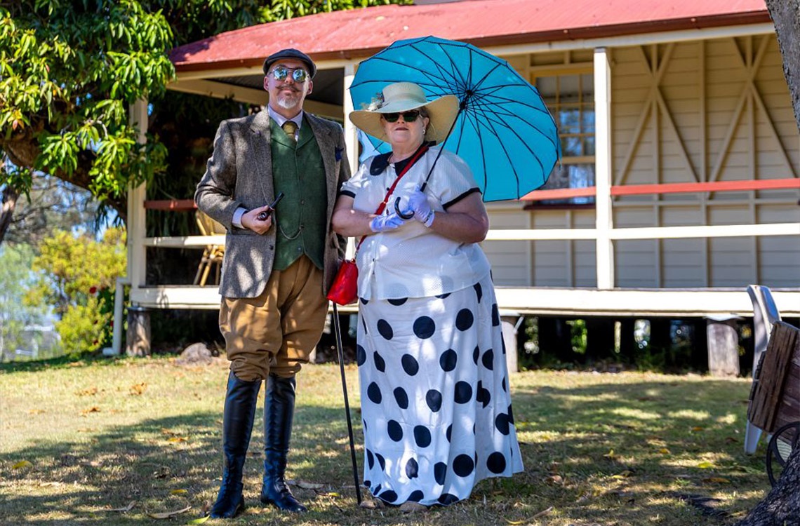 Couple dressed in 1930s dress holding a blue umbrella in Mayes Cottage garden