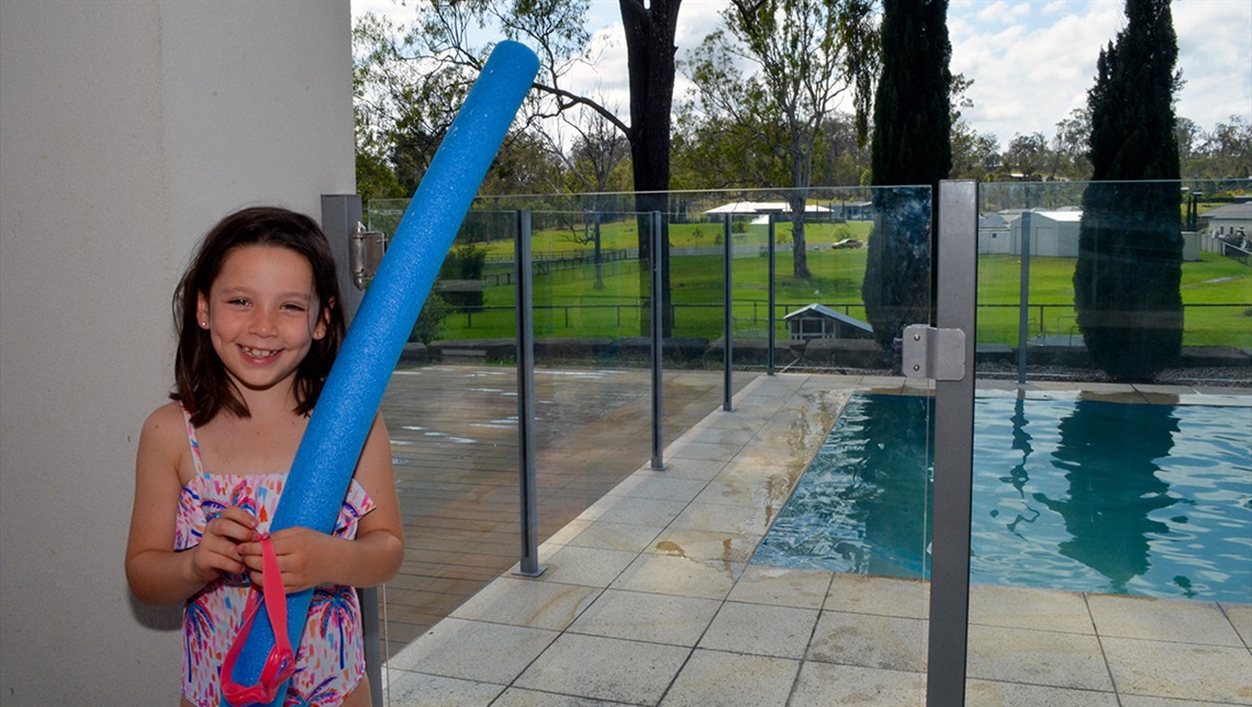A girl holding a blue pool noodle and pink goggles standing in from on a pool fenced with glass panels