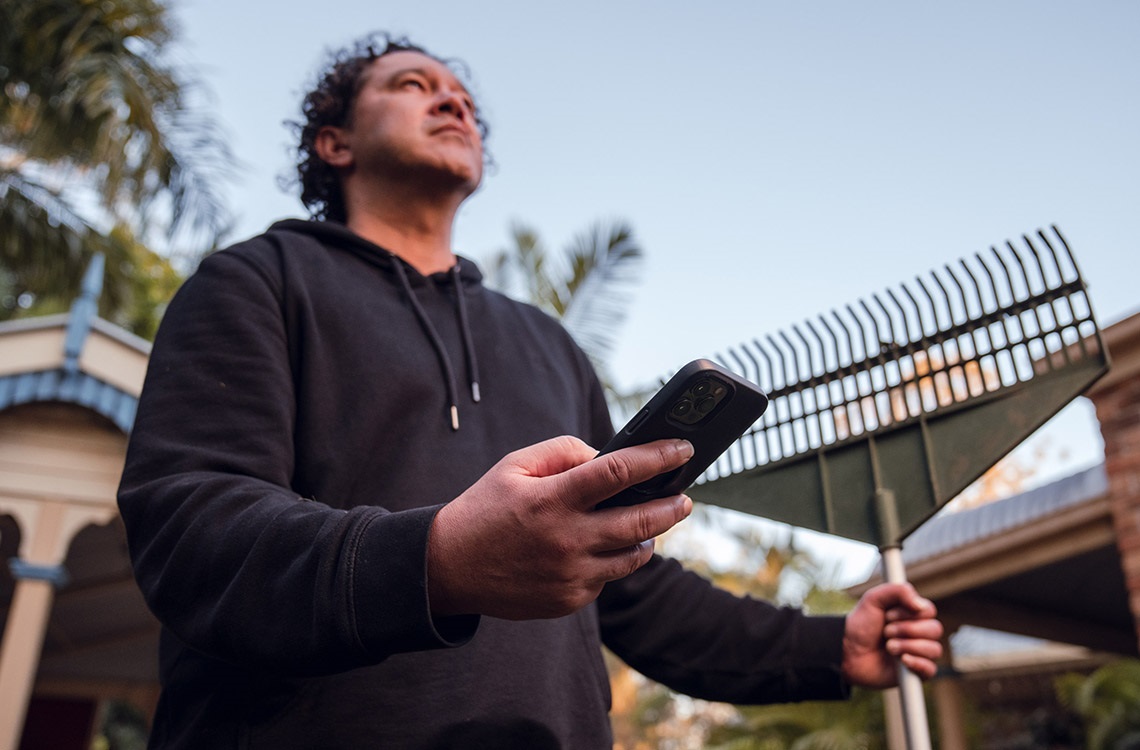 Man in yard holding rake and phone and looking at the weather in the sky